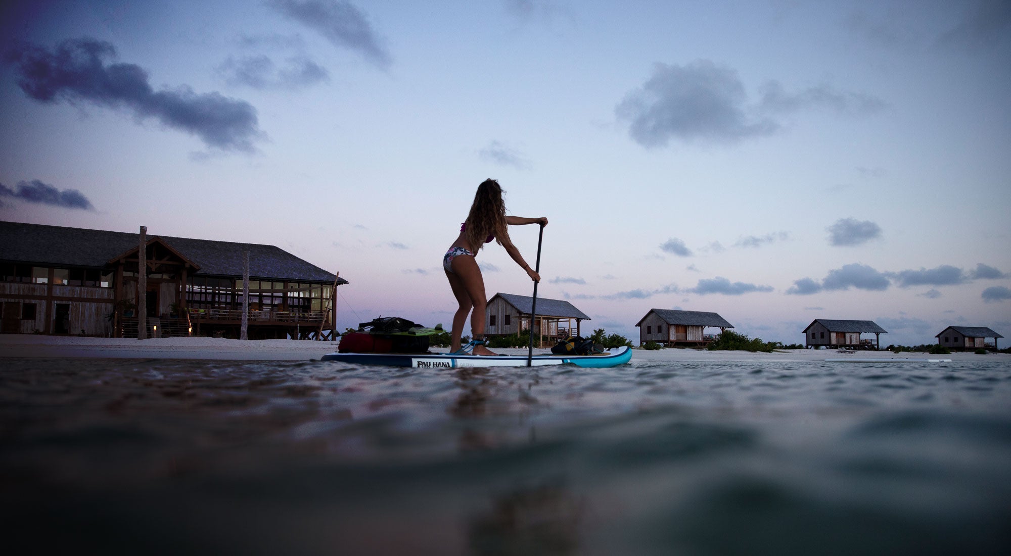 a woman paddling an inflatable paddleboard past a resort in the barbuda at dusk