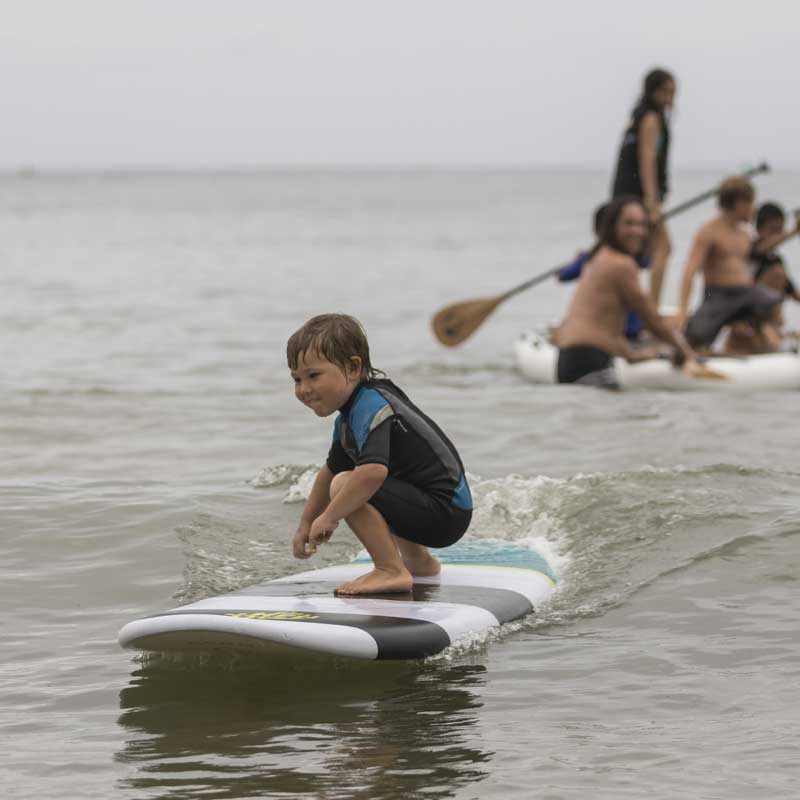A kid surfing a paddleboard on a wave with a group of adults in the background