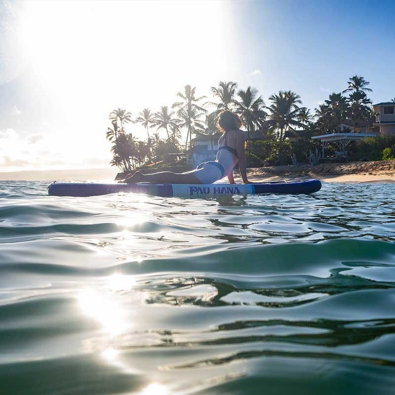 A woman doing yoga on an inflatable paddleboard with beach homes and palm trees in the background