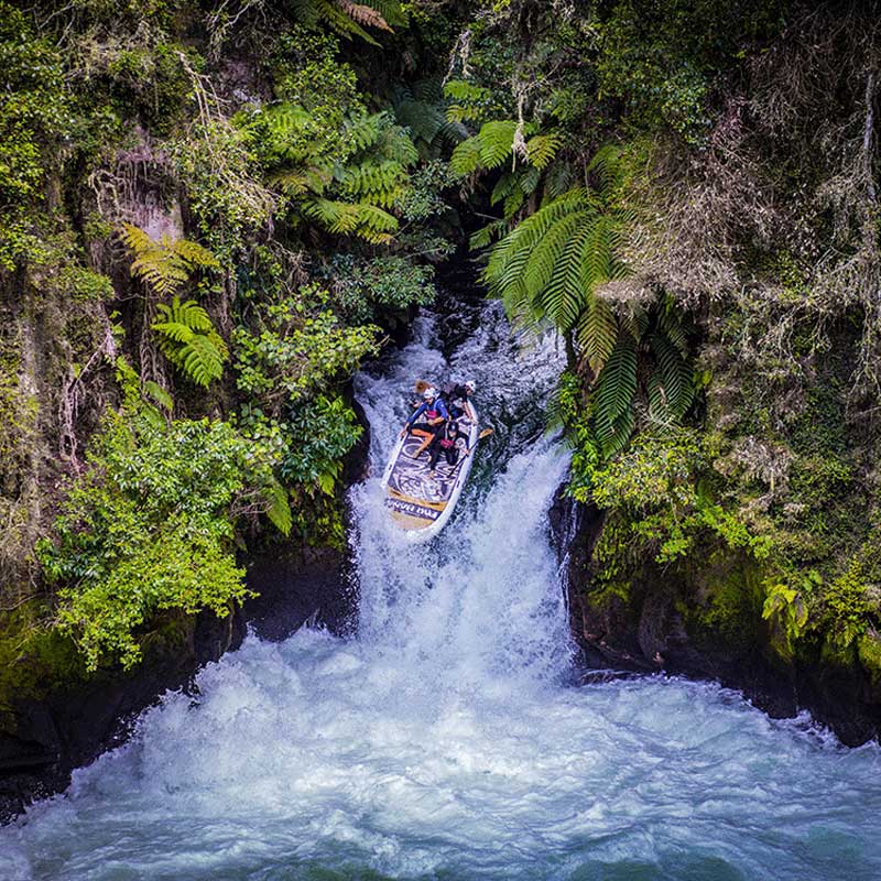 a giant inflatable paddleboard going over a waterfall surrounded by new zealand native bush