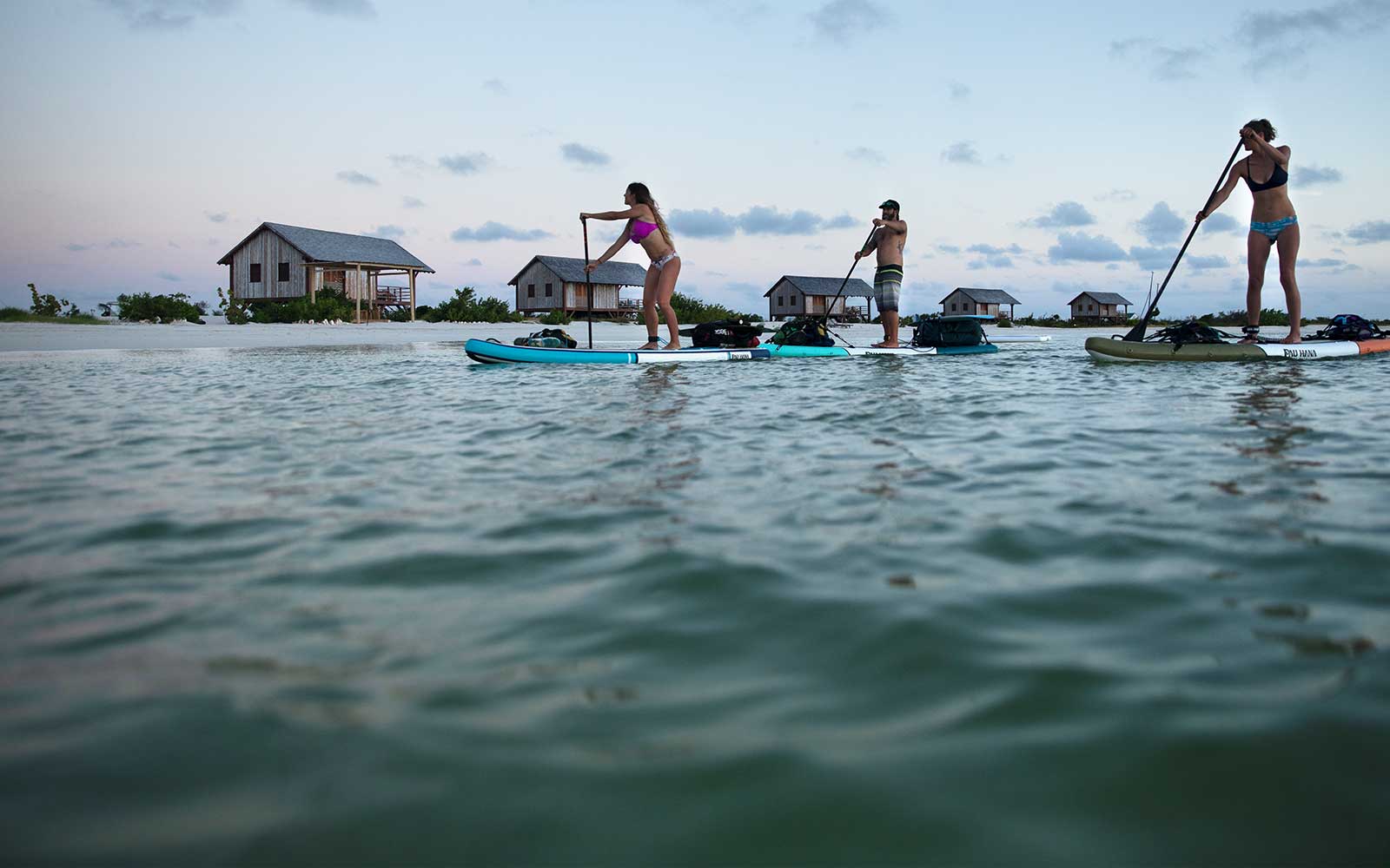 three stand up paddleboarders paddling past ocean front huts in barbuda