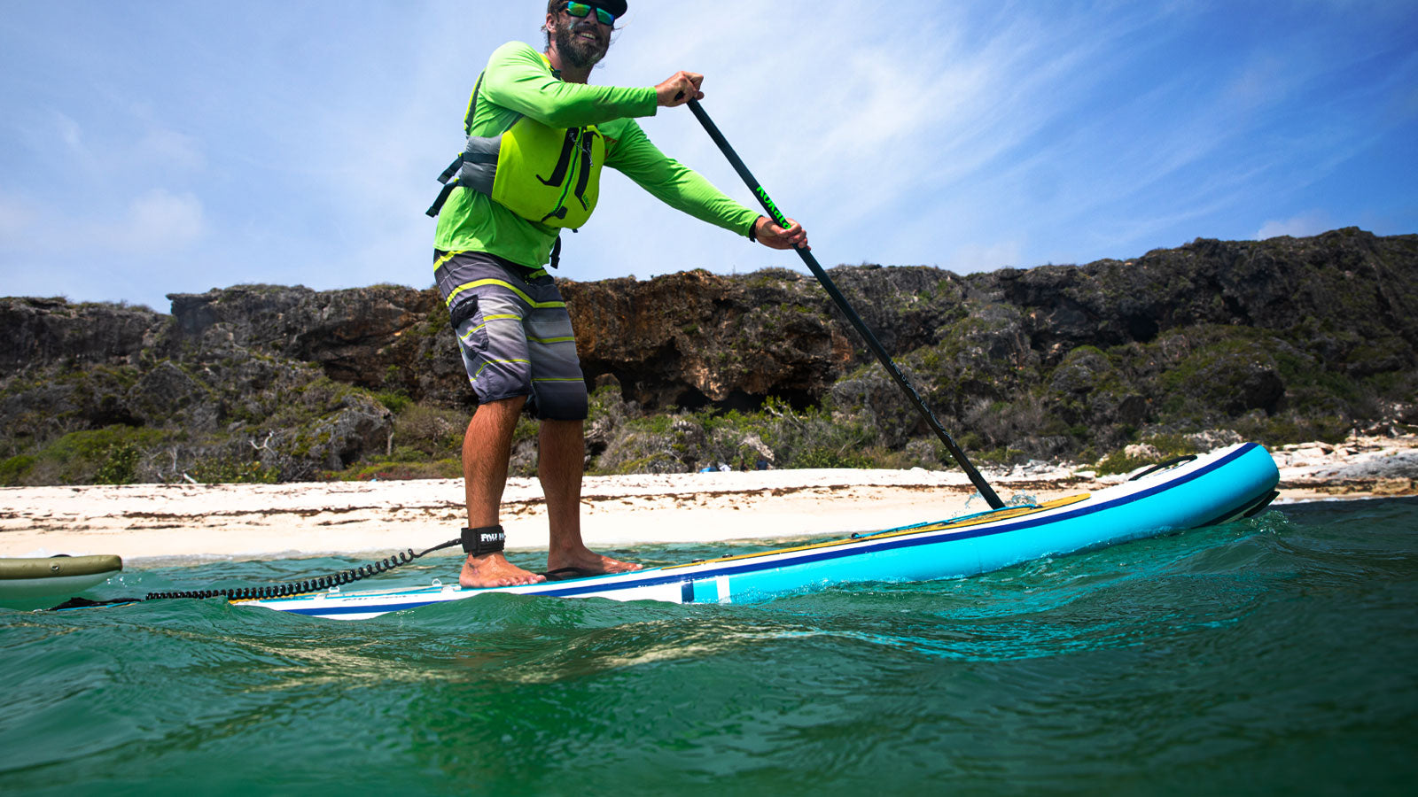 A happy man paddleboarding infront of a beach with cliffs in the background