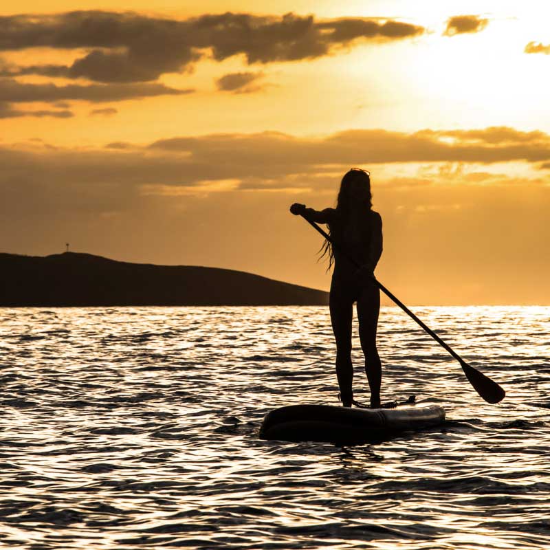 a woman stand up paddleboarding at sunset with an outcrop of land in the background