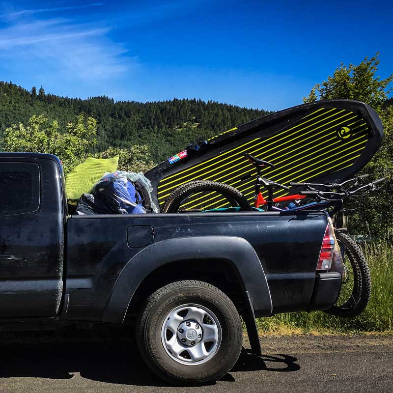 A stand up paddleboard in the back of a pickup truck with a bike and gear next to it