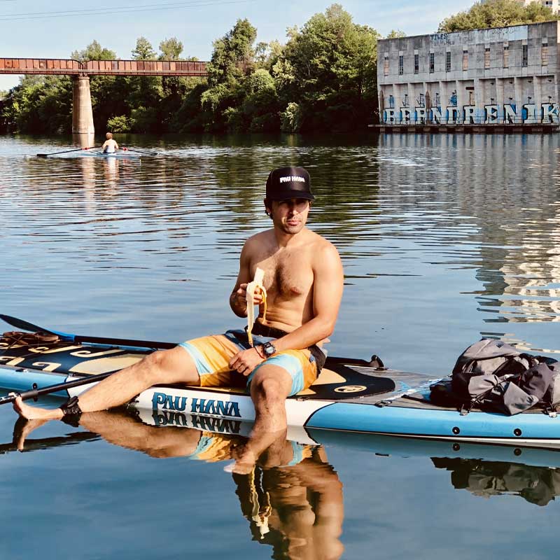 a guy sat eating a banana on the pau hana endurance xl paddleboard with an old building and bridge in the background