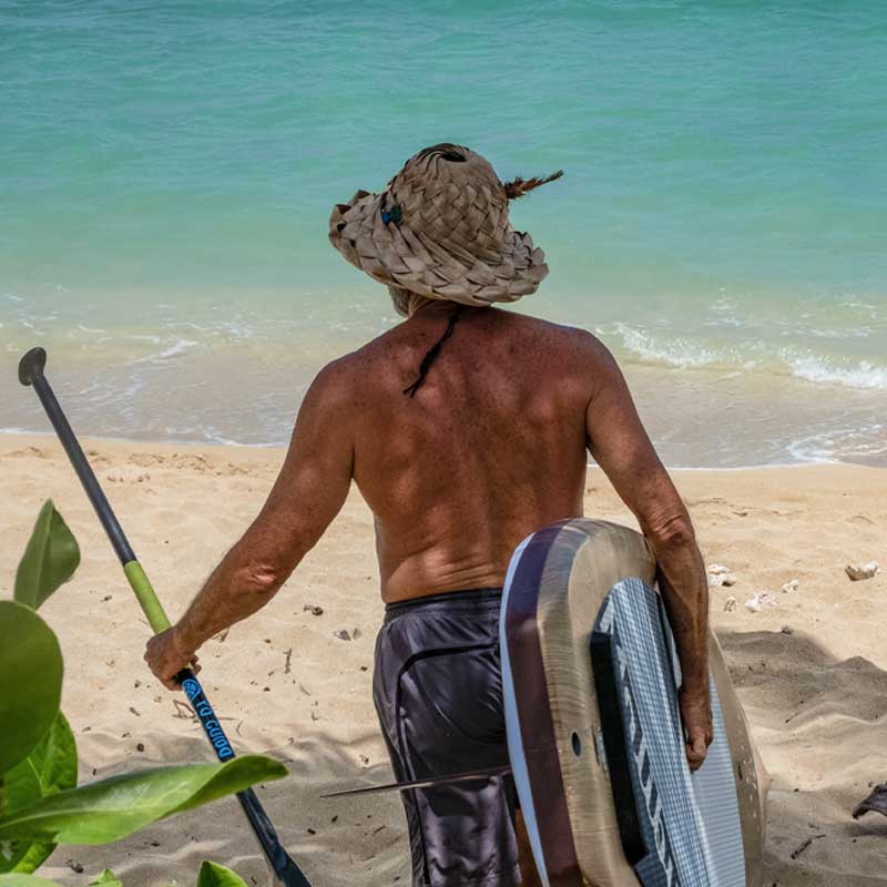 a guy carrying the malibu tour paddleboard onto the beach wearing a straw hat