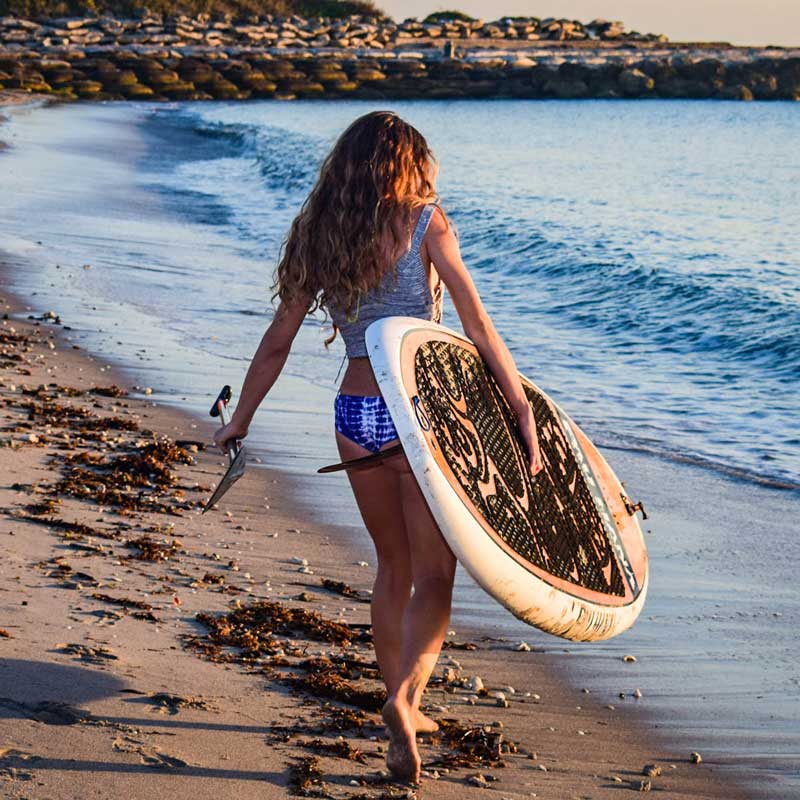 a woman carrying the oahu paddleboard at sunset down a beach